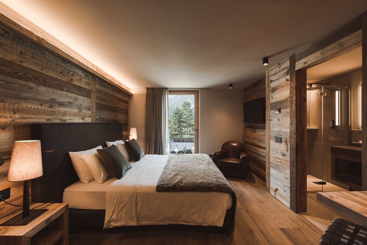 Un dormitorio moderno con una cama grande, paredes de madera, un televisor y vista a árboles.