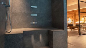 A modern bathroom with a built-in shower, a bench, and a glass door leading to a room with a bed and a lamp. - Hotel Alù (Bormio)