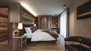 A modern bedroom with a large bed, wooden headboard, bedside tables, a TV, and a chair. - Hotel Alù (Bormio)
