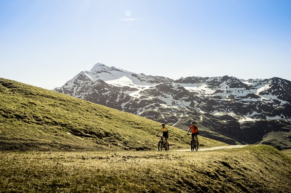 A mountain biking trail with two cyclists, surrounded by snow-capped mountains and a clear sky. - Hotel Alù (Bormio)