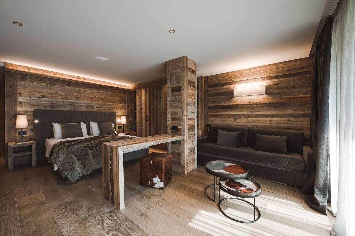 Un dormitorio moderno con un panel de madera decorativo, una cama con cabecera, una mesita de noche, un sofá y una pequeña mesa redonda con un adorno.