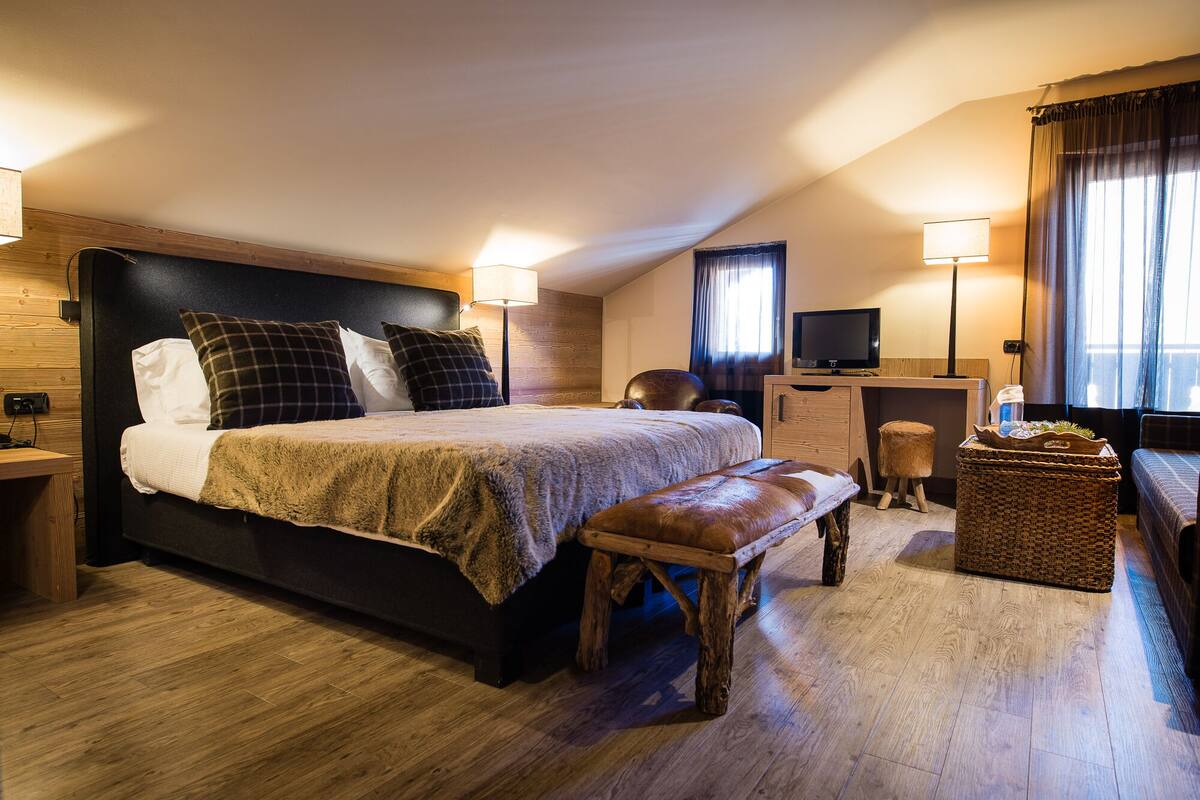 Un dormitorio con una cama grande, un banco de madera, un escritorio con televisor y una ventana con cortinas.