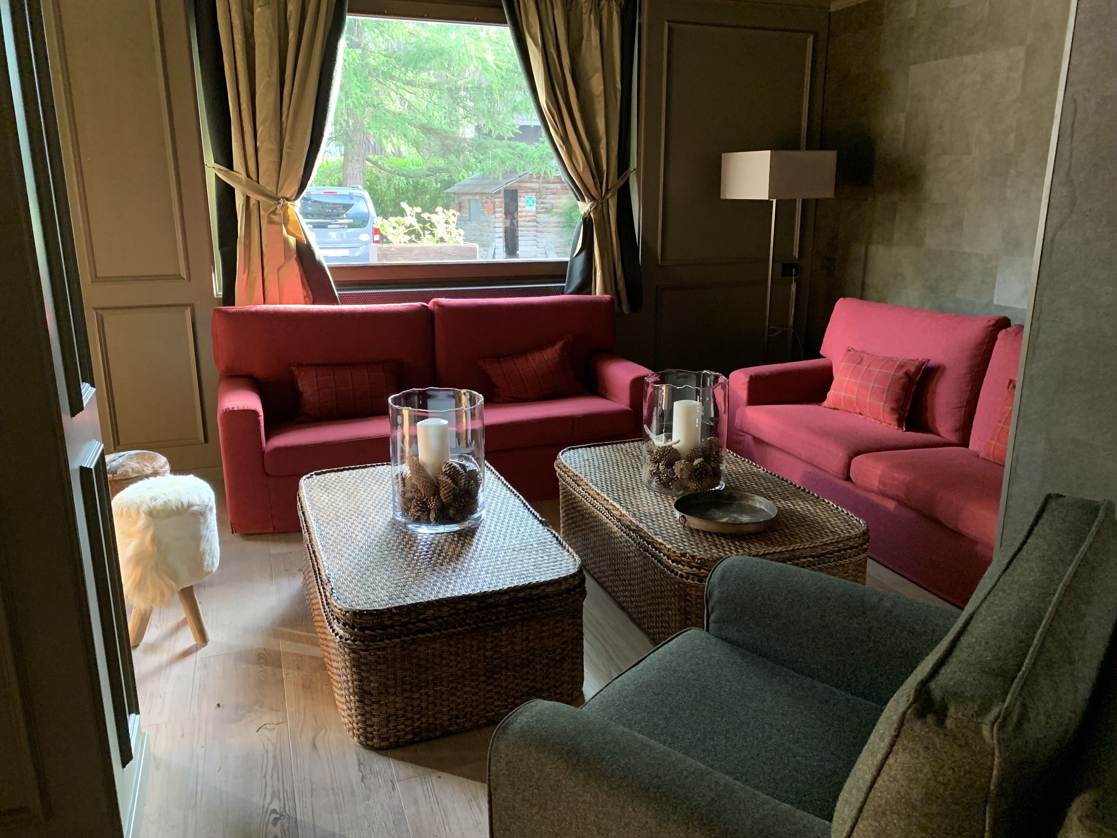 Una sala de estar acogedora con un sofá rojo, dos otomanas de mimbre, un sillón verde y una ventana con vistas a un coche y vegetación.