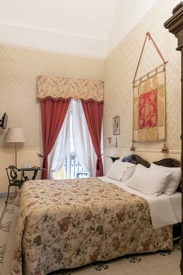 Une chambre à coucher avec une couvre-lit à motifs floraux, un lit recouvert de draps blancs, une table de chevet avec une lampe et une fenêtre ornée de rideaux rouges.