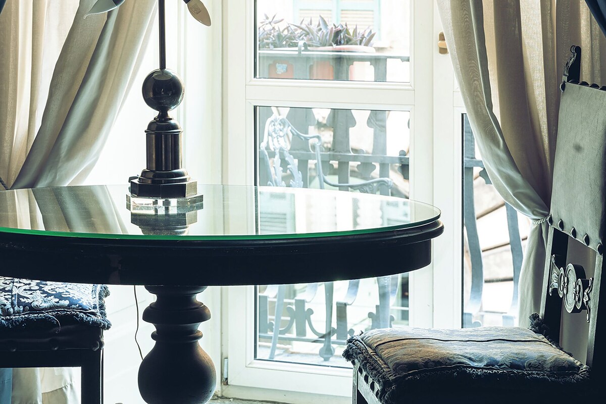 Una habitación con una mesa de vidrio, una lámpara y vistas a un edificio a través de una ventana con cortinas.