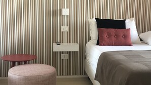 A hotel room with a striped wall, a bed with white and red pillows, a small round table, and a pink ottoman. - Mia Hotel Numana (Numana)
