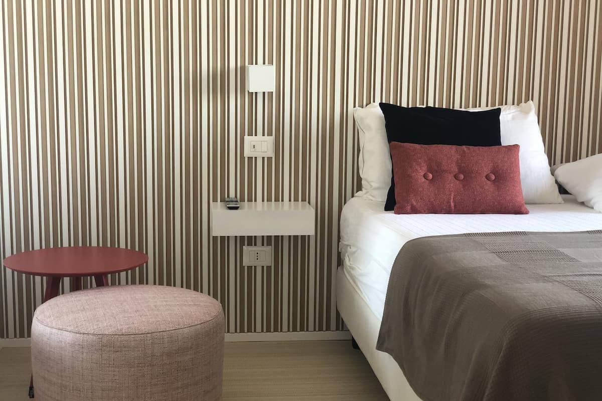 A hotel room with a striped wall, a bed with white and red pillows, a small round table, and a pink ottoman.