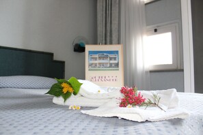 A neatly made bed with a framed picture of a hotel, a bouquet of flowers, and a folded towel on a patterned bedspread.