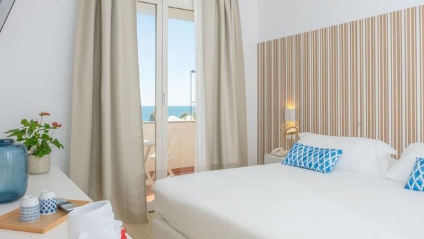 A hotel room with a bed, a desk with a vase and a cup, and a view of the sea through a sliding door. - Mia Hotel Numana (Numana)