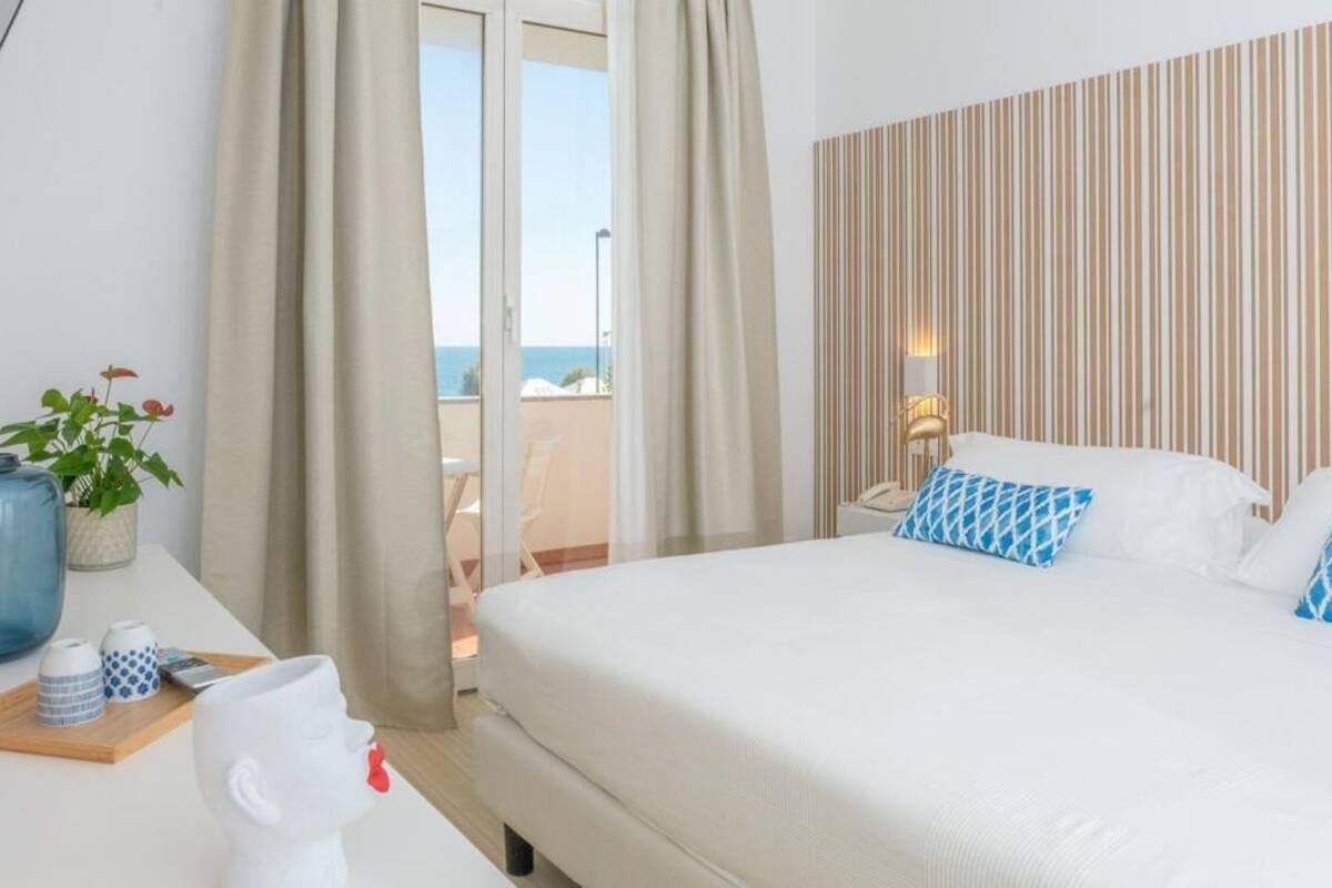 A hotel room with a bed, a desk with a vase and a cup, and a view of the sea through a sliding door.