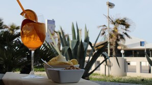 A glass of drink with a straw and a slice of fruit, a bowl of chips, and a small bowl of nuts on a table with a view of palm trees and a building in the background. - Mia Hotel Numana (Numana)