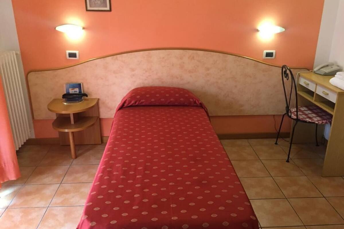 A single bed with a red bedspread, a bedside table with a lamp, a chair, and a small desk with a telephone.