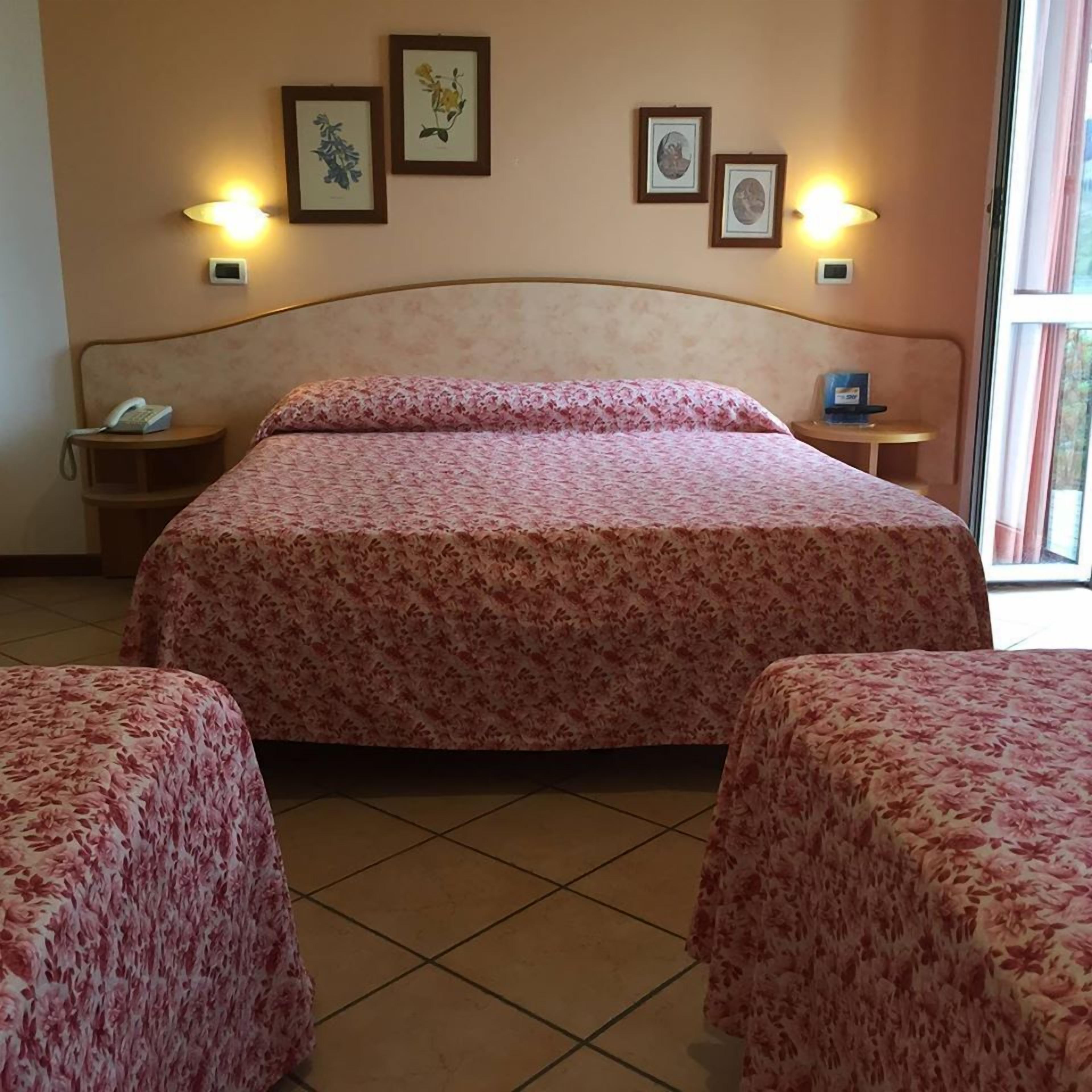Ein Hotelzimmer mit einem großen Bett, zwei Einzelbetten, einem Nachttisch mit Telefon und gerahmten Pflanzenbildern an der Wand.