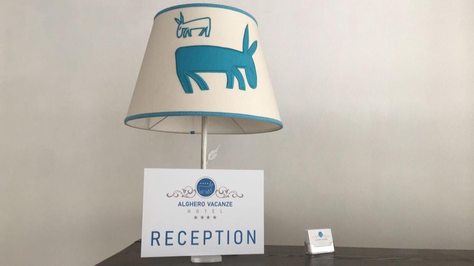 A reception desk with a lamp featuring a blue donkey design, a sign indicating the hotel name 'ALGHERO VACANZE HOTEL', and a reception sign.