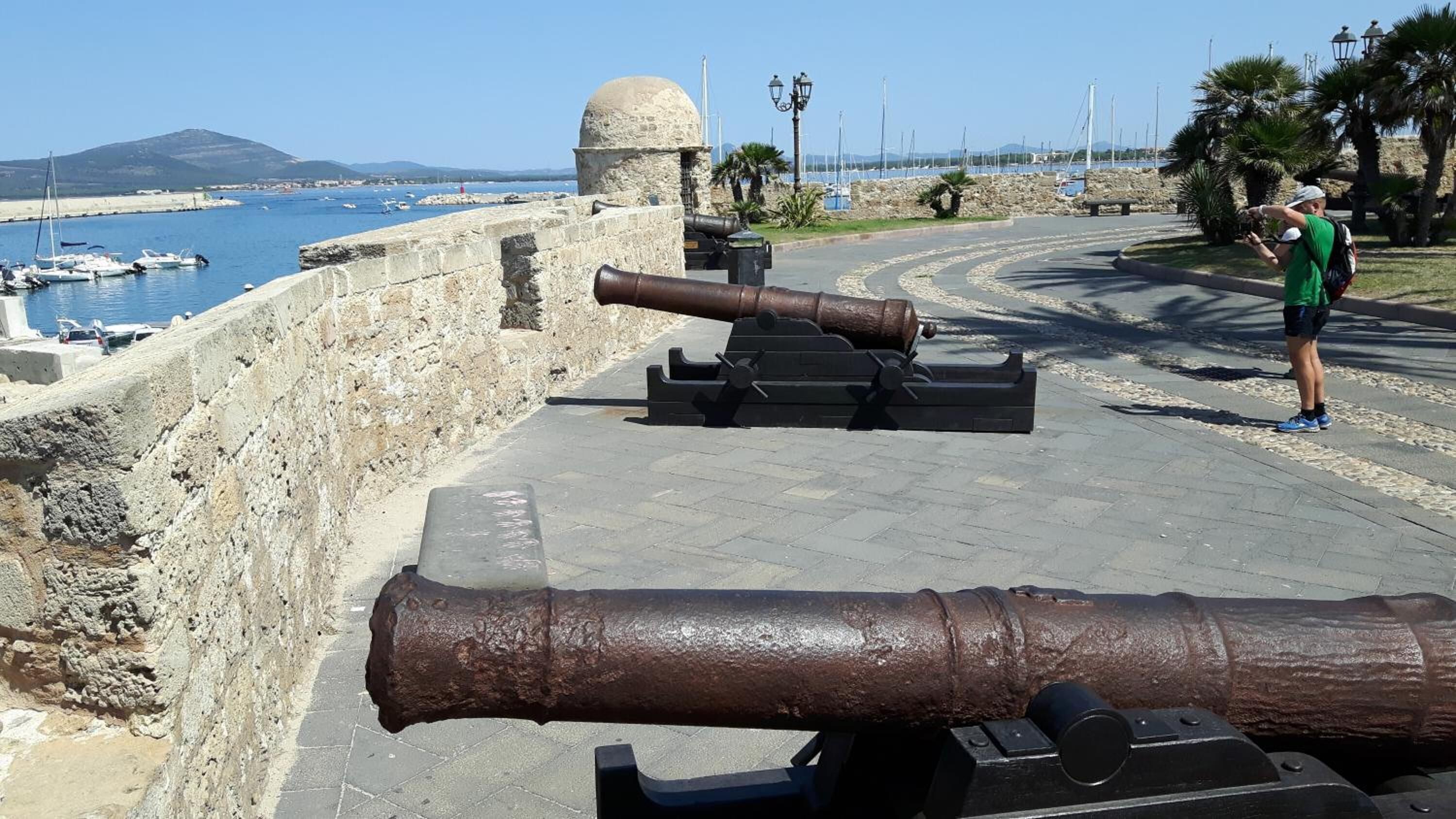 A coastal fortification with cannons, a stone wall, and a clear view of the sea and boats.