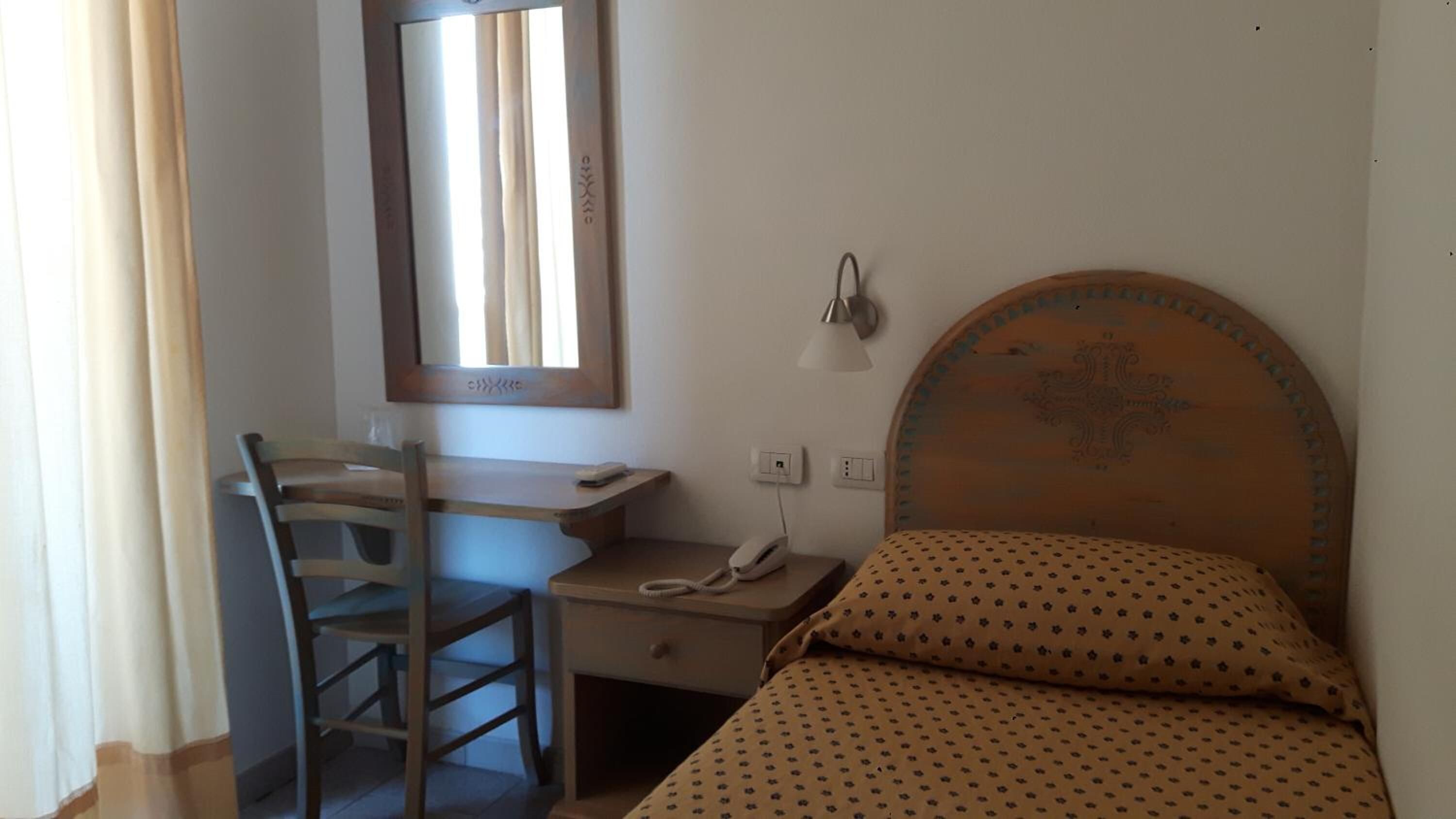A small room with a wooden desk, a chair, a bed with a patterned pillow, a wall-mounted lamp, and a window with curtains.