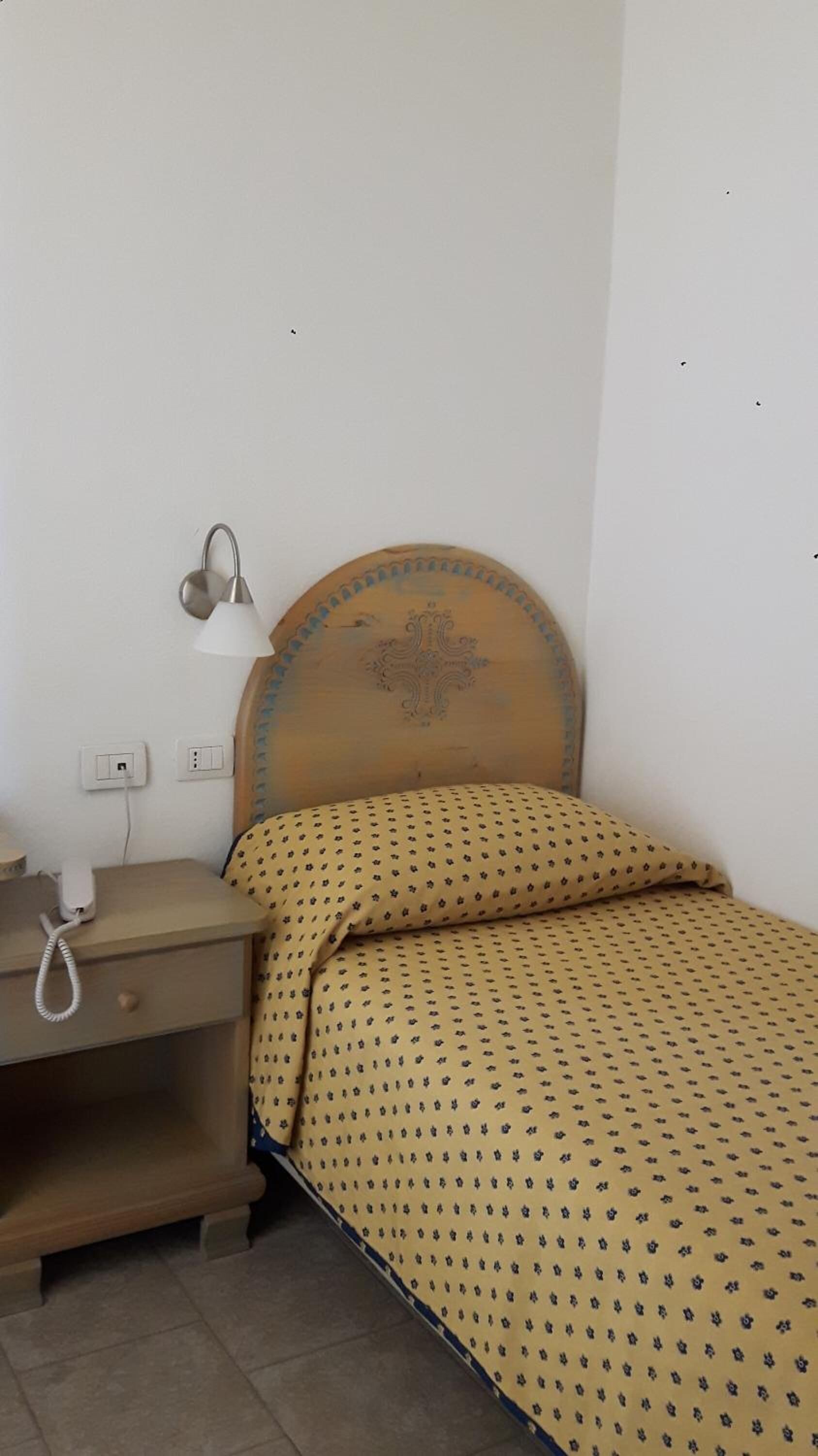 Ein kleiner, schlichter Raum mit einem hölzernen Bett, einem Nachttisch mit Lampe und einem Telefon.
