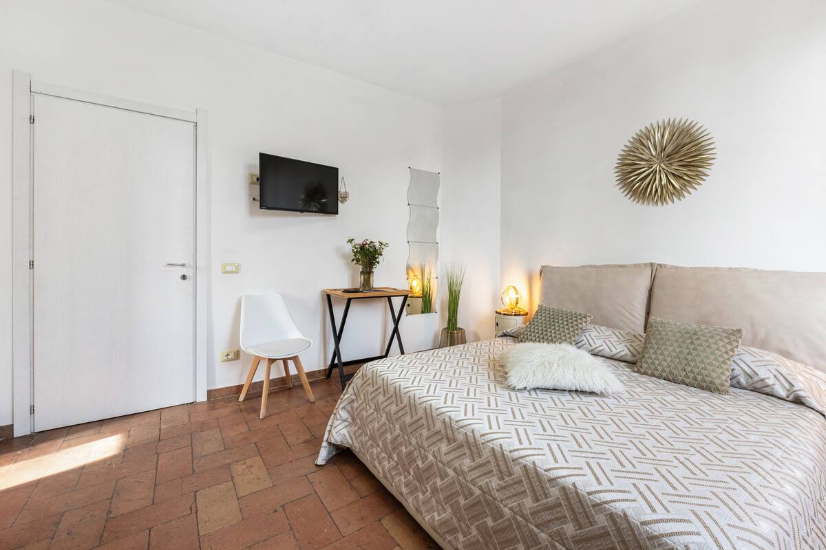 A bedroom with a bed, a TV mounted on the wall, a chair, a small table, and a decorative wall piece.