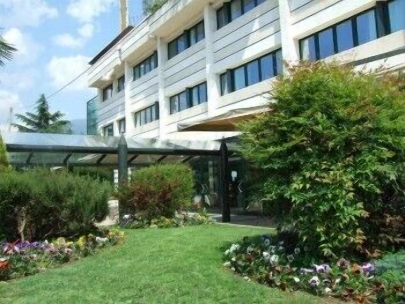 Un bâtiment moderne doté d’une entrée en verre et d’un jardin bien entretenu, où l’on trouve une variété de plantes et de fleurs.