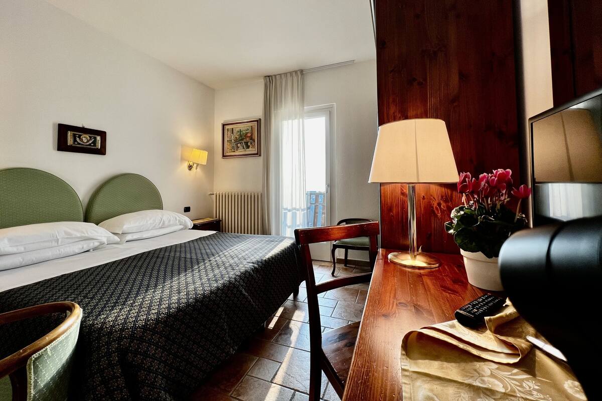 Une chambre d’hôtel avec un lit, un bureau doté d’une lampe, une chaise, une plante en pot et un tableau au mur.