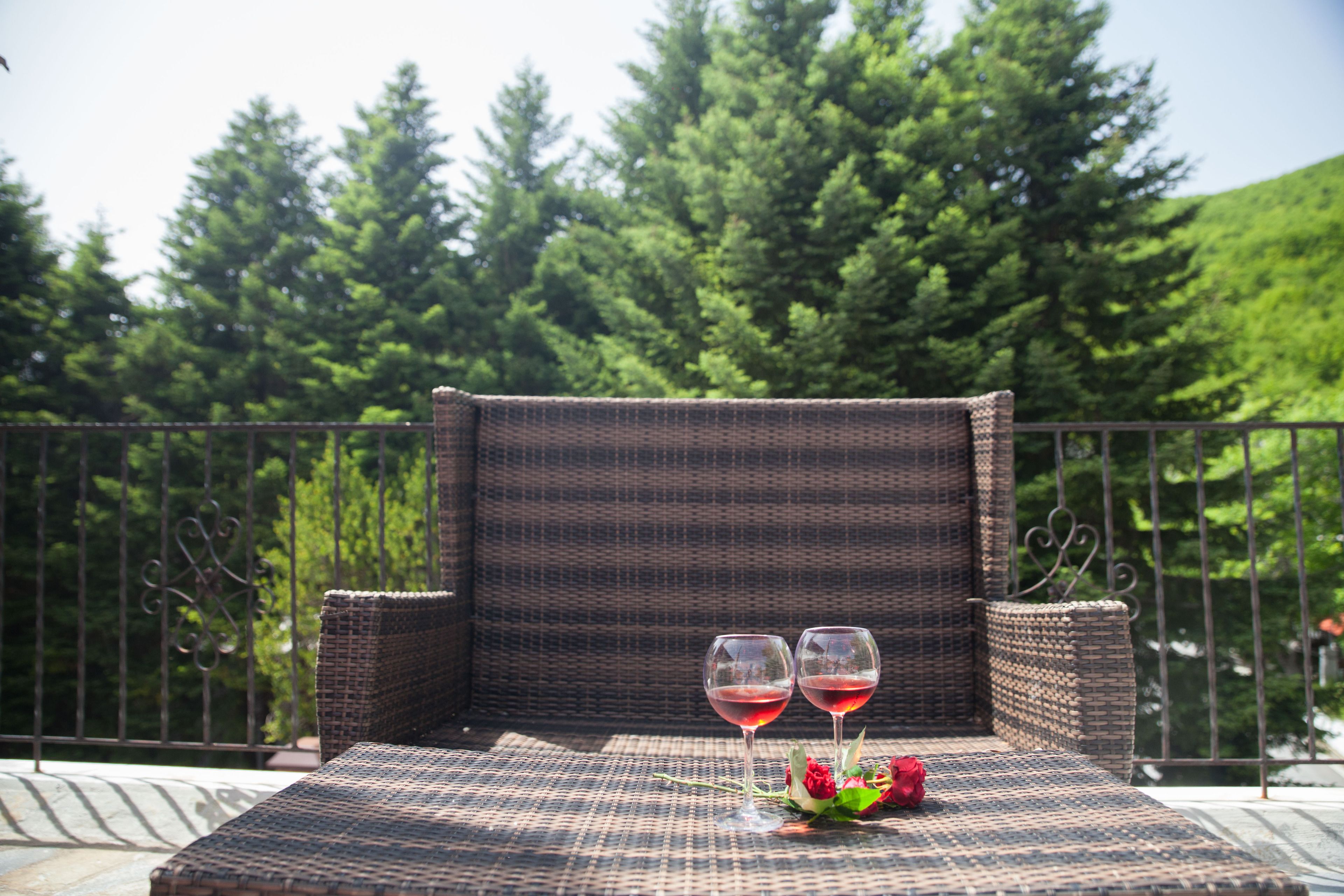 Una silla de mimbre con una mesa, dos copas de vino y un ramo de flores en un balcón con vistas a árboles.