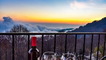 Un balcón con una botella de vino y dos copas, con vistas a un paisaje montañoso al atardecer.