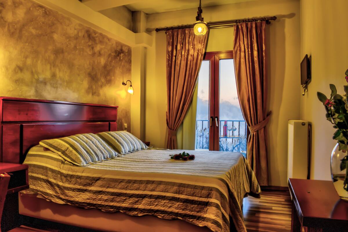 A hotel room with a large bed, a wooden headboard, a bedside table, a nightstand, a potted plant, and a view of a balcony with curtains.