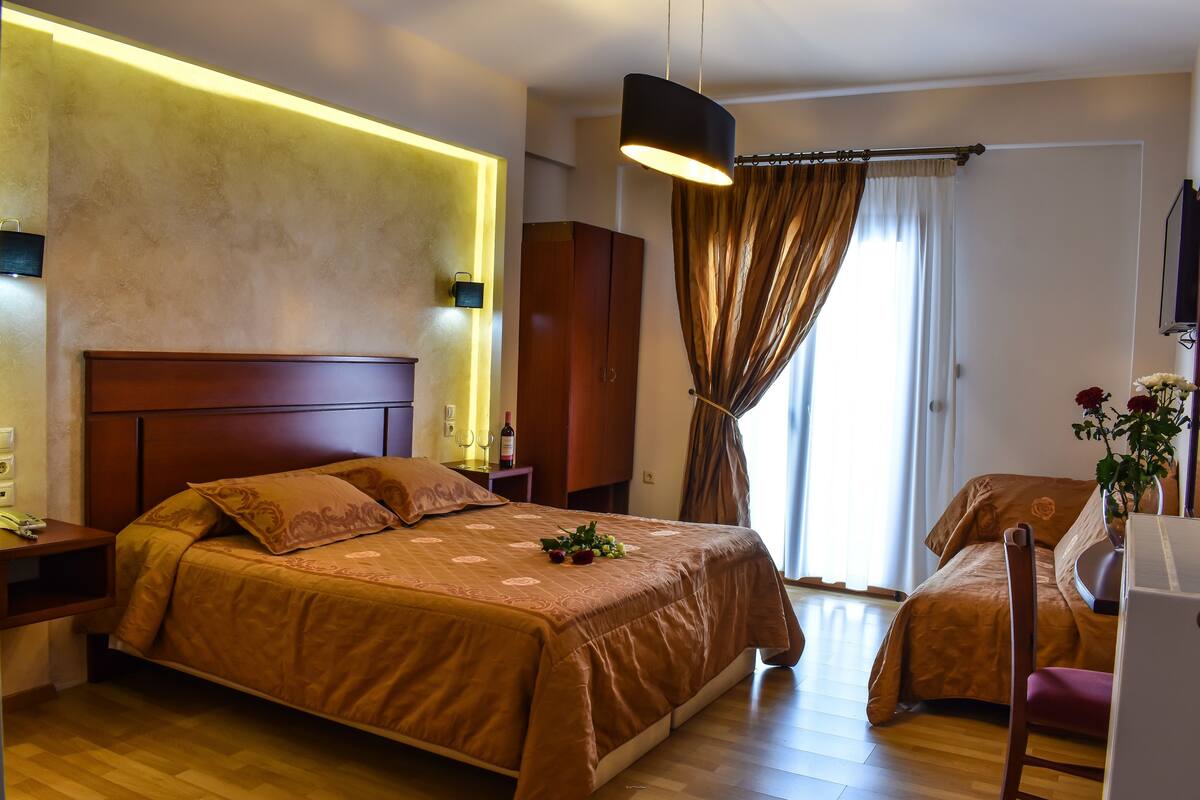 A hotel room with a large bed, bedside tables, a desk with a chair, a potted plant, and a window with curtains.