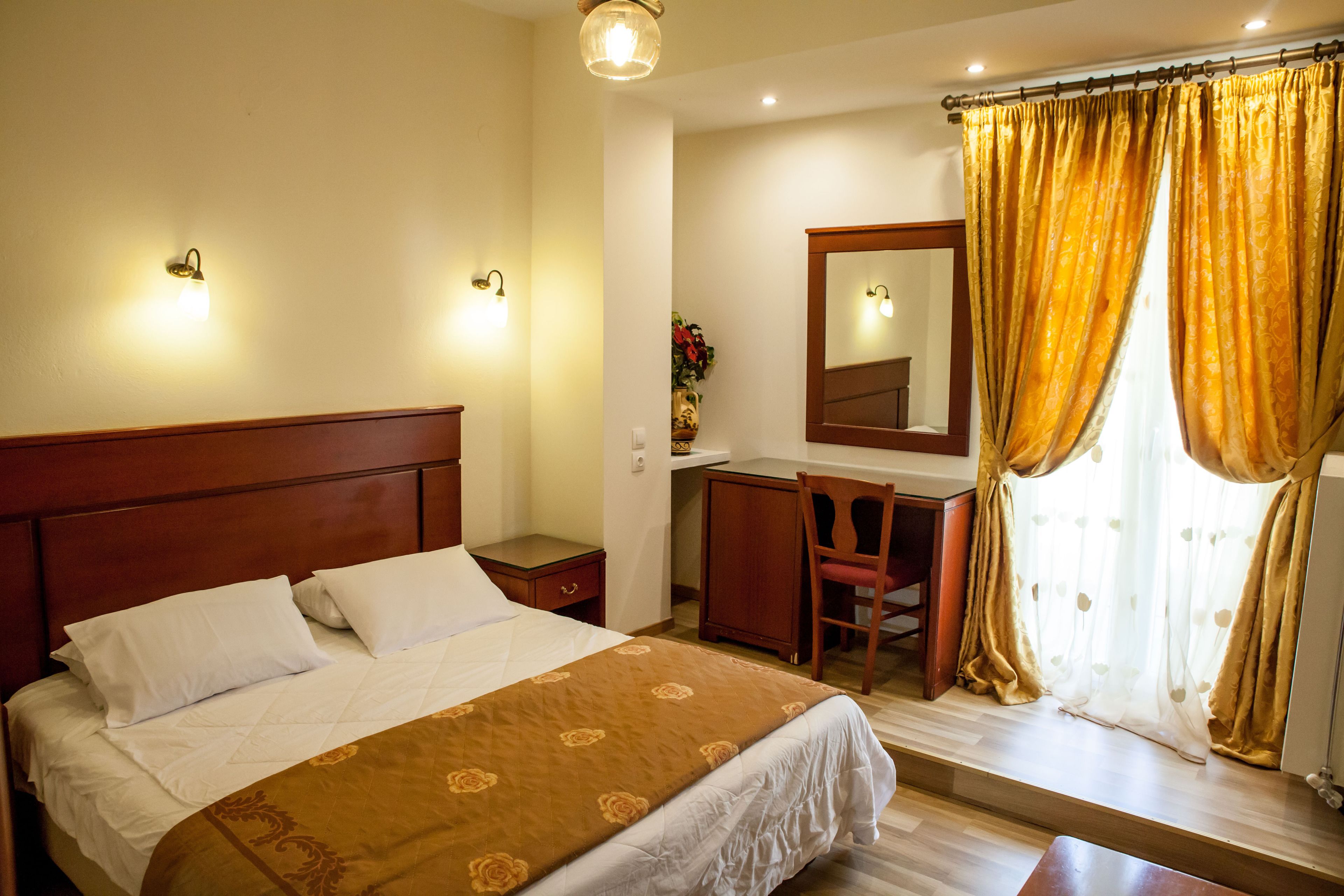 A hotel room with a large bed, a wooden nightstand, a desk with a chair, a mirror, and a window with curtains.