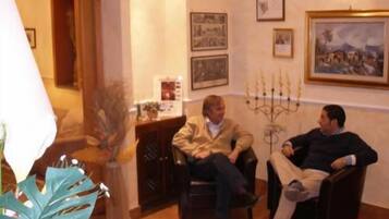 Deux hommes sont assis dans une pièce où se trouvent une lampe, un chandelier et des tableaux encadrés accrochés au mur.
