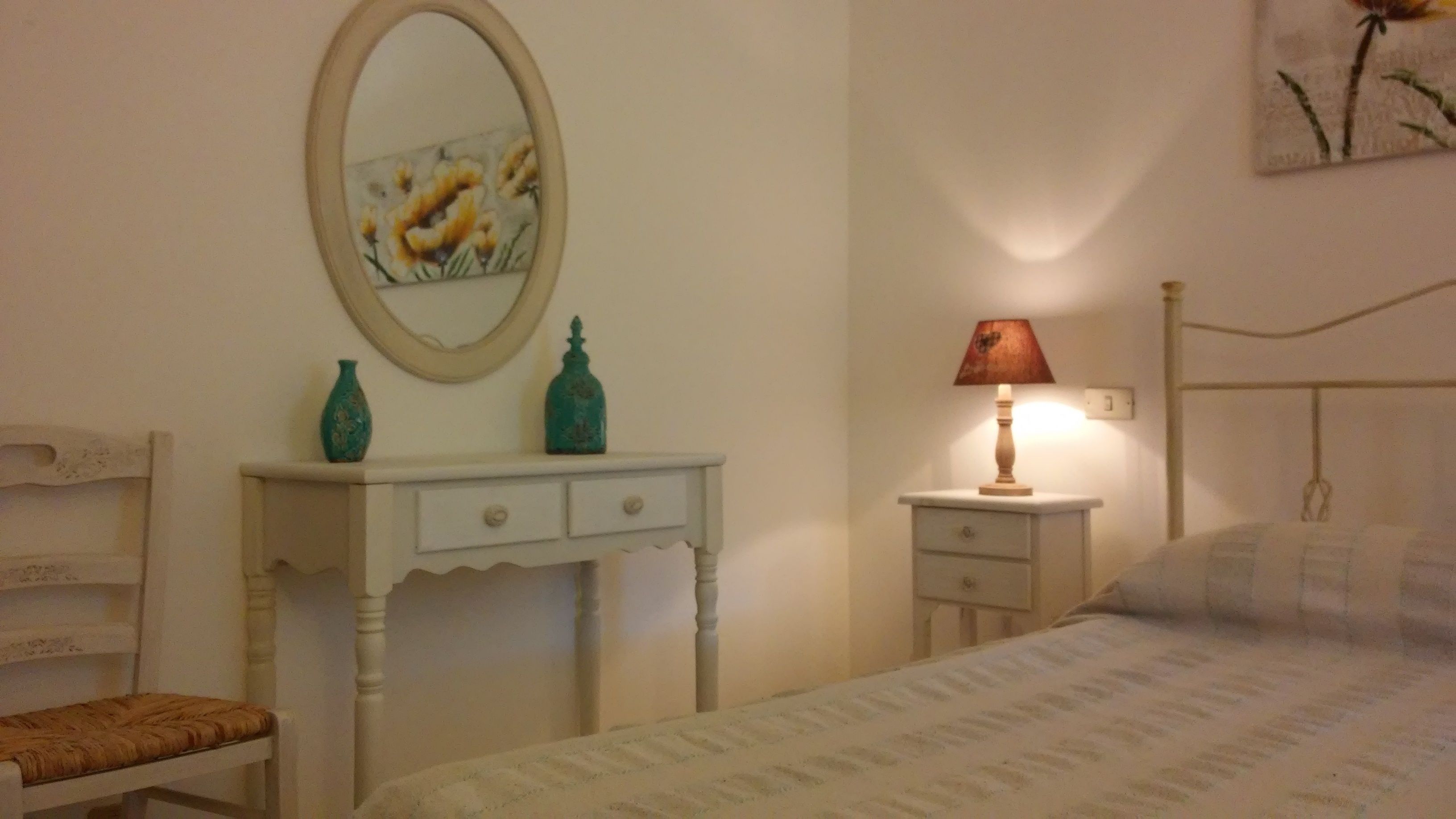 A bedroom with a bed, a nightstand with a lamp, a dresser with two decorative bottles, a mirror, and a painting on the wall.