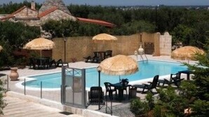 Zona de piscina con sillones de descanso y sombrillas, rodeada de vegetación y un edificio de piedra con tejado rojo.