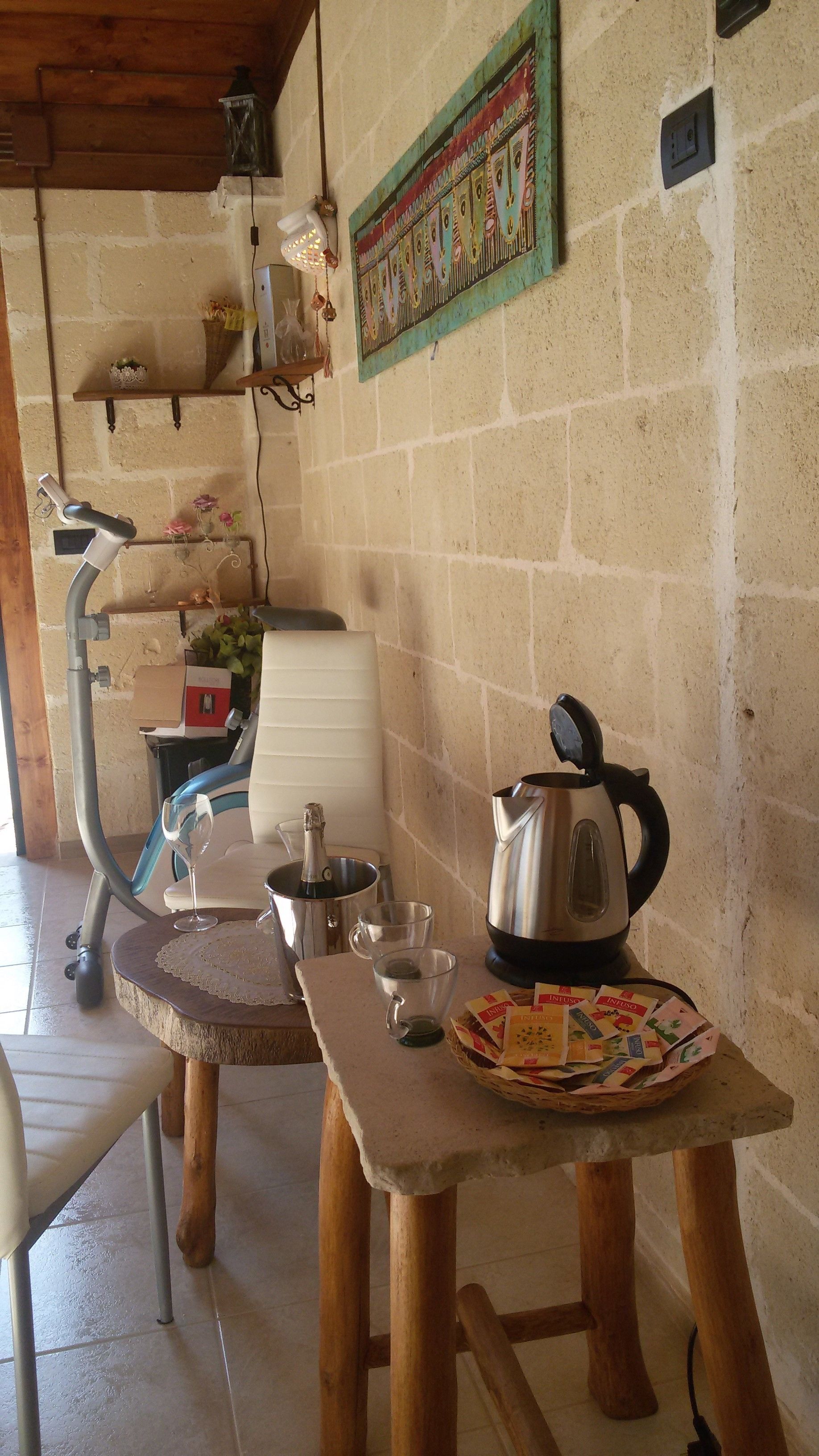 A rustic kitchen with a stone wall, a wooden table with a kettle, a coffee pot, and a basket of tea bags.