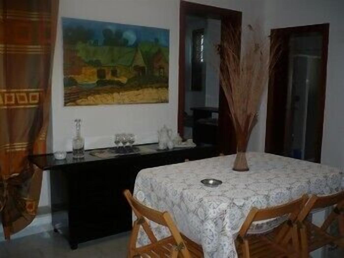 Un comedor con una mesa preparada para cuatro personas, un cuadro en la pared y un jarrón con plantas secas.