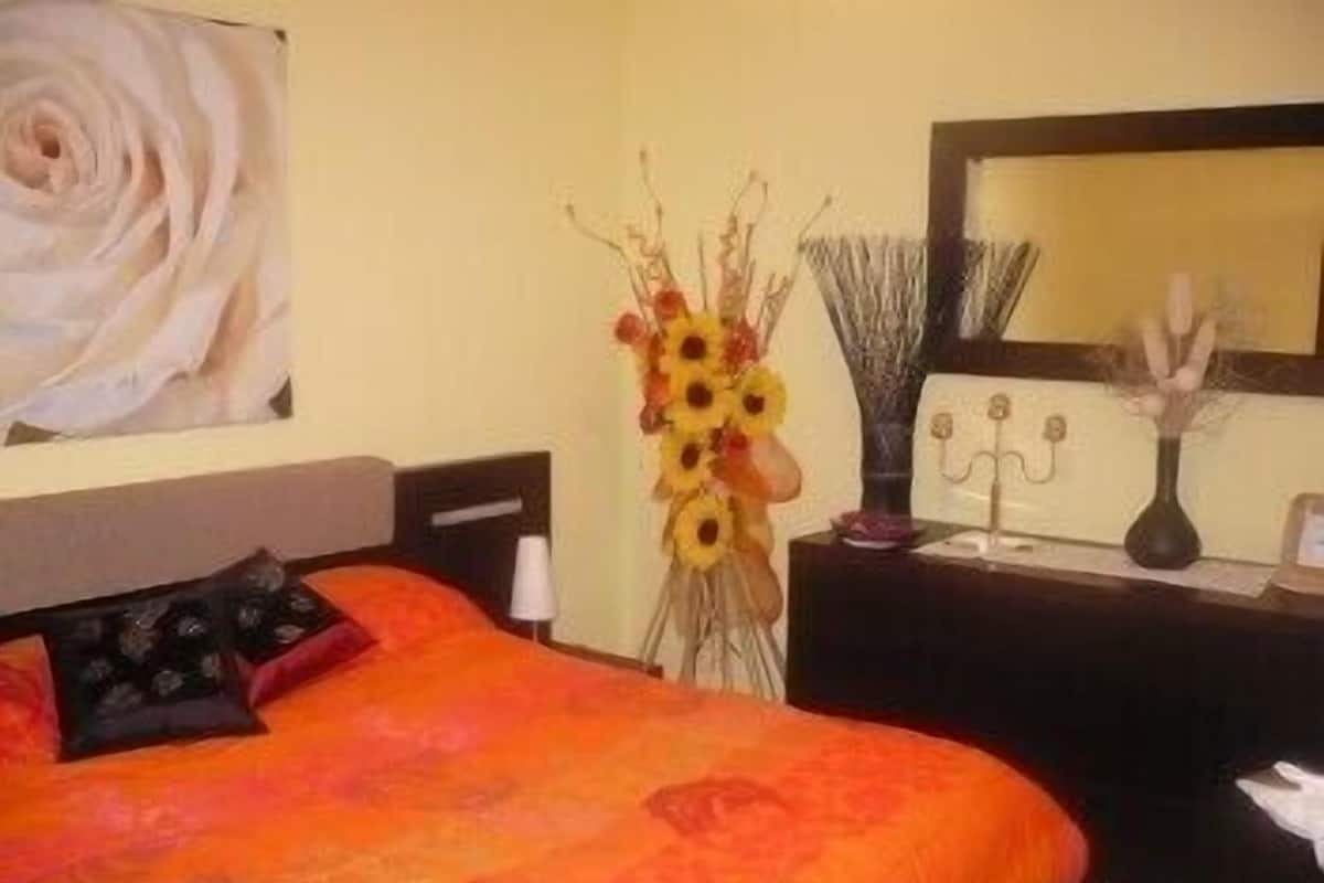 A bedroom with a large bed, a nightstand, a vase with sunflowers, and a wall-mounted mirror.