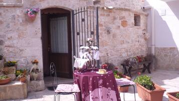 Un espace extérieur aménagé pour les repas, avec une table dressée pour deux personnes, entouré de plantes en pot et bordé d’un bâtiment en pierre au style rustique.