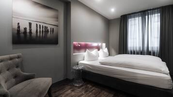 Una habitación de hotel moderna con una cama grande, un sillón capitonado, una mesita de noche y una obra de arte colgada en la pared.