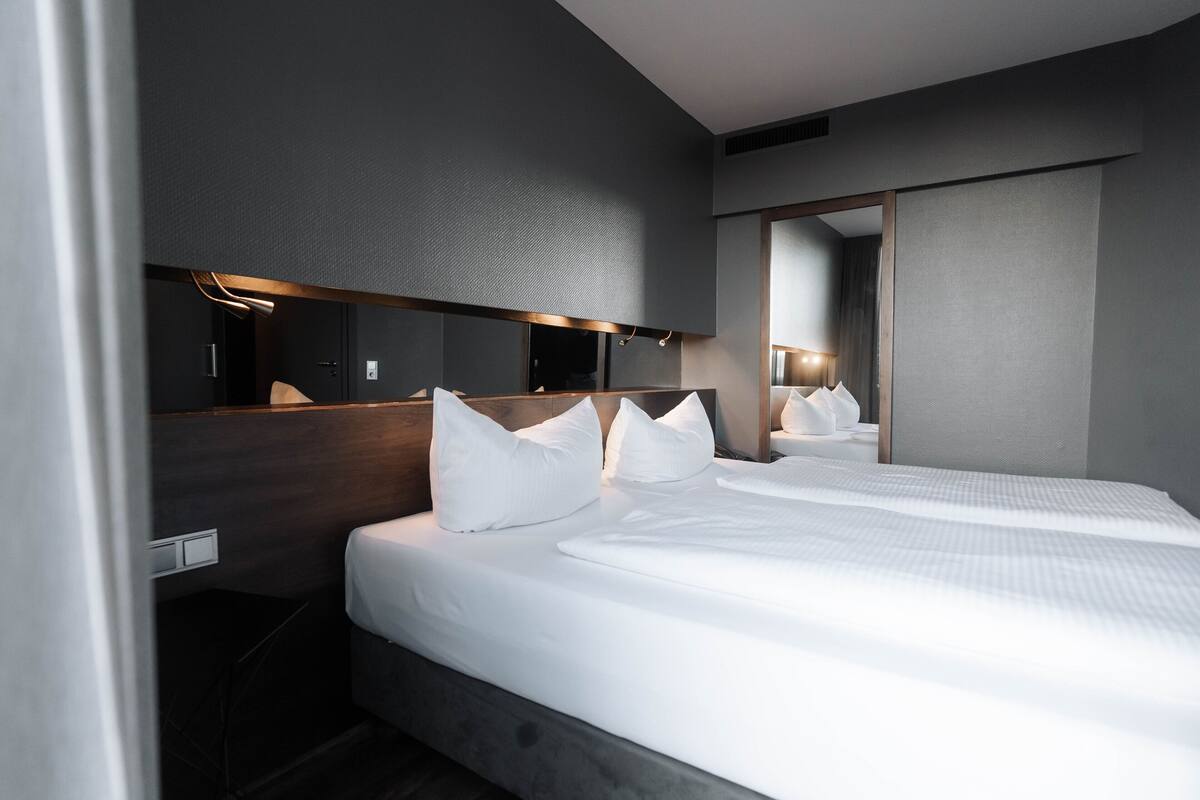 A modern hotel room with a large bed, a bedside table, a mirror, and a wall-mounted light fixture.