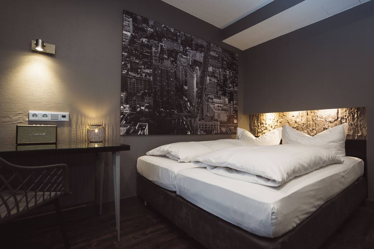 A modern hotel room with a large cityscape artwork on the wall, a desk with a lamp, and a neatly made bed.