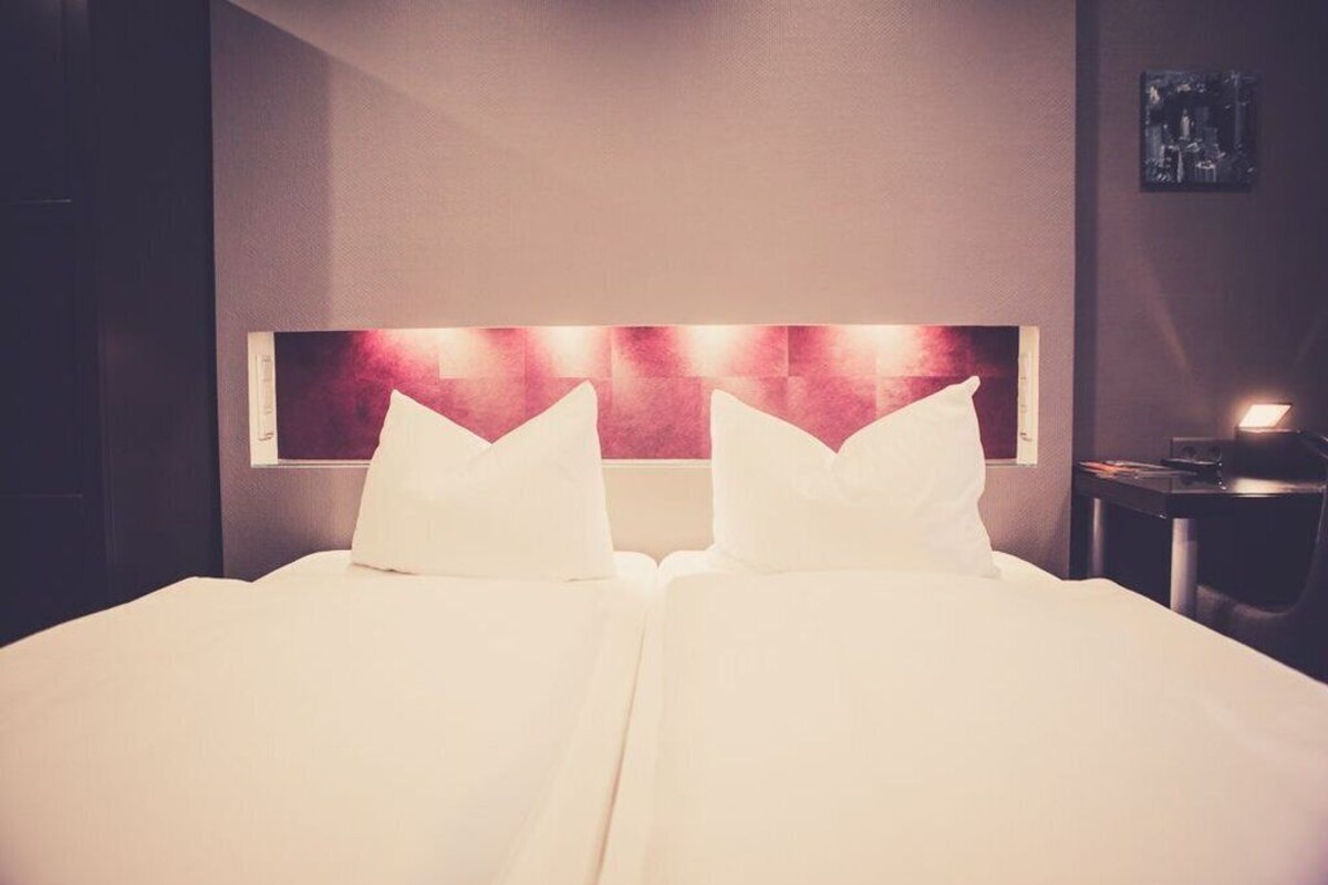 A hotel room with a large bed, a headboard with a red panel, and a bedside table with a lamp.
