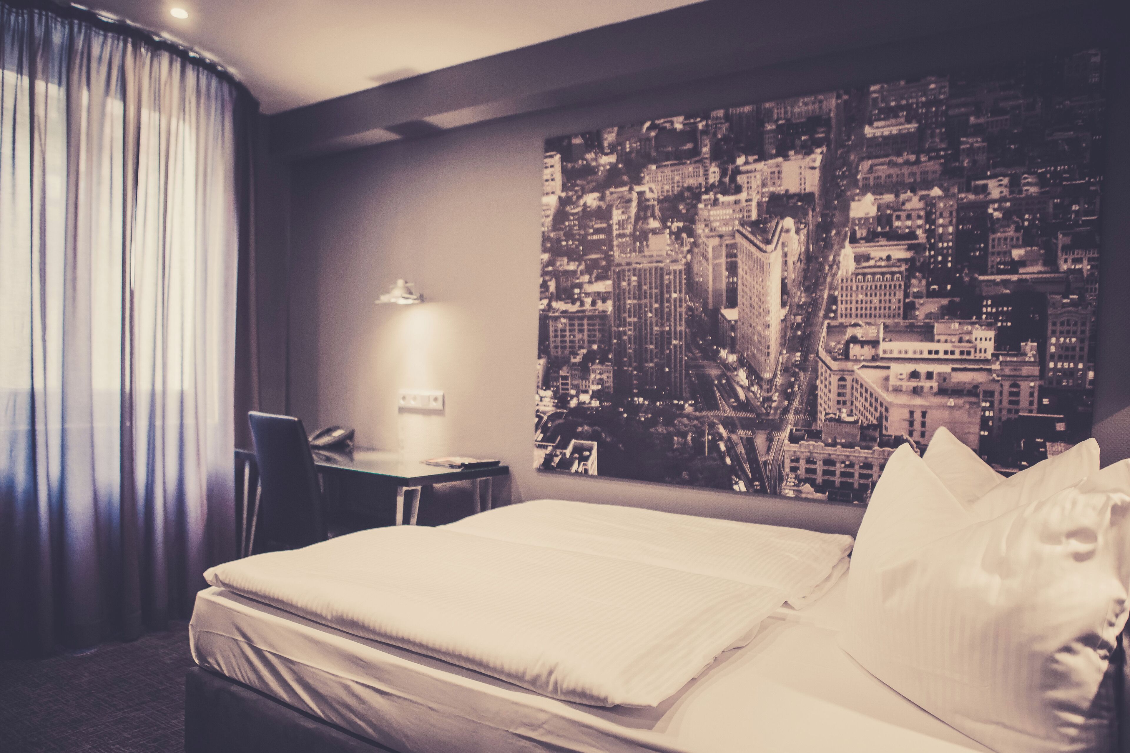 A hotel room with a bed, a desk, a chair, a lamp, and a cityscape view.