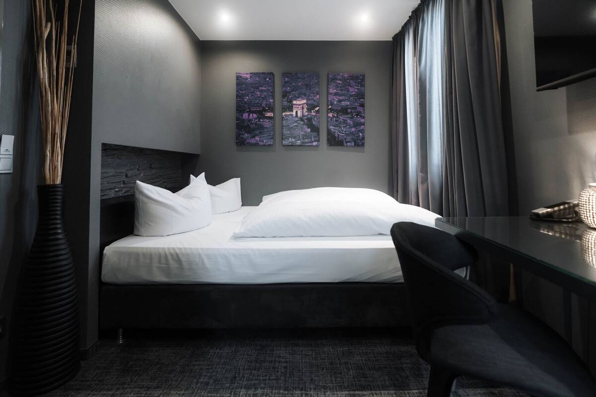 A modern hotel room with a bed, a desk, and a chair. There are three paintings on the wall, a vase, and a lamp on the desk.