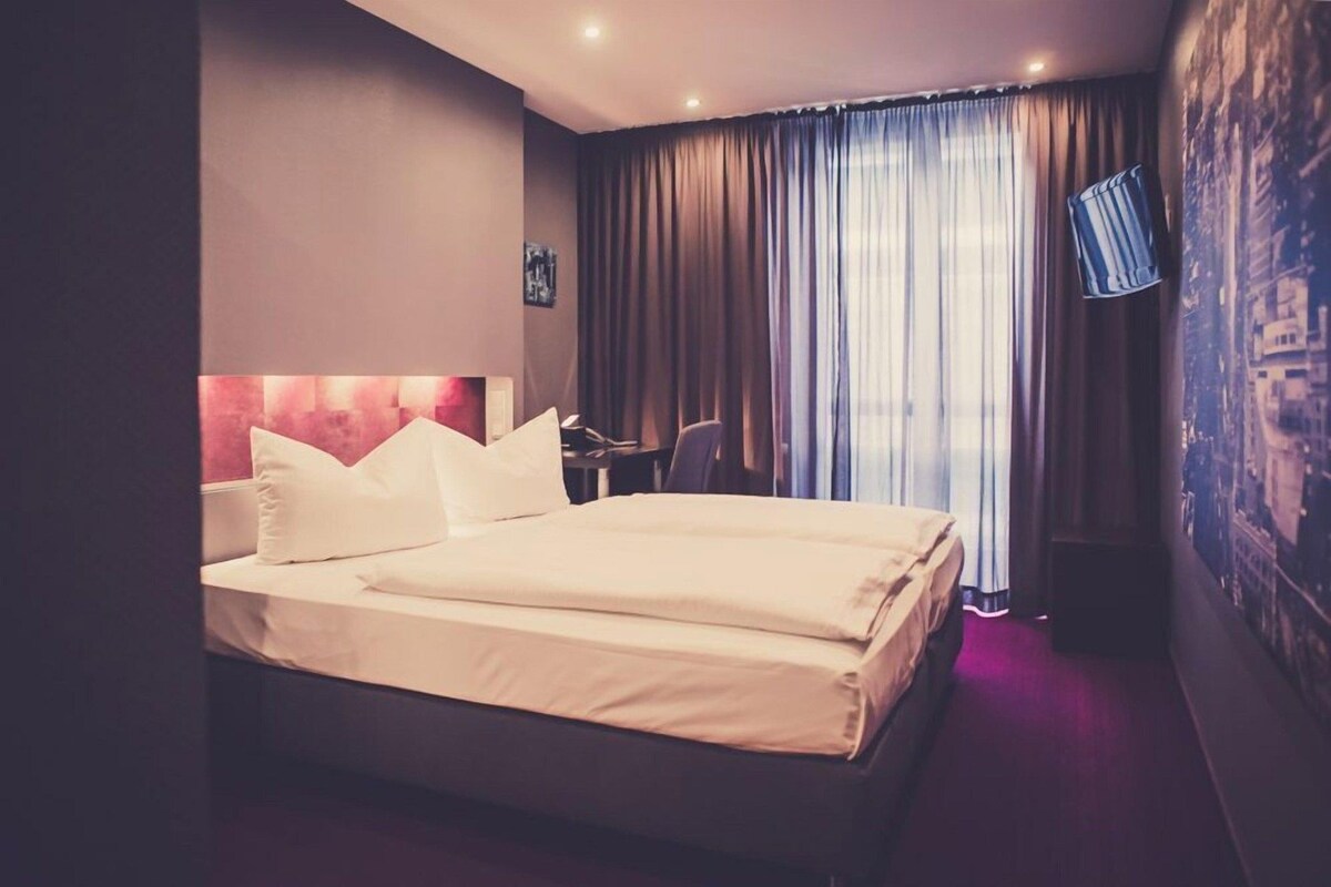 A hotel room with a large bed, a desk, a chair, and a TV mounted on the wall.