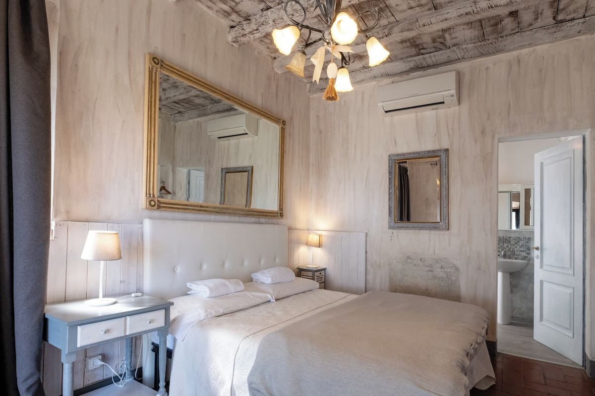 A bedroom with a large bed, bedside tables with lamps, a ceiling light fixture, a large mirror, and an air conditioning unit.
