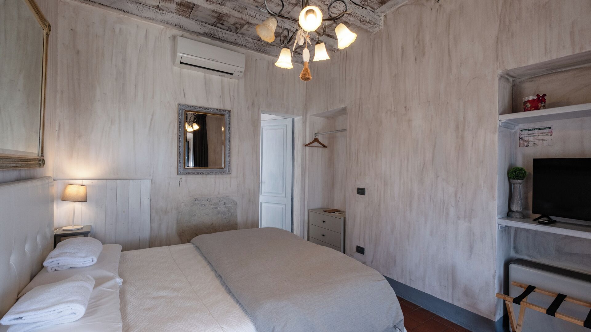 Une chambre à coucher comprenant un lit, une table de chevet, un miroir, un luminaire suspendu, une armoire et un téléviseur posé sur une étagère.