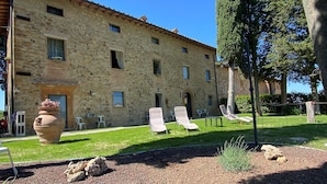 A stone building with a tiled roof, outdoor seating area with lounge chairs, and a gravel path.