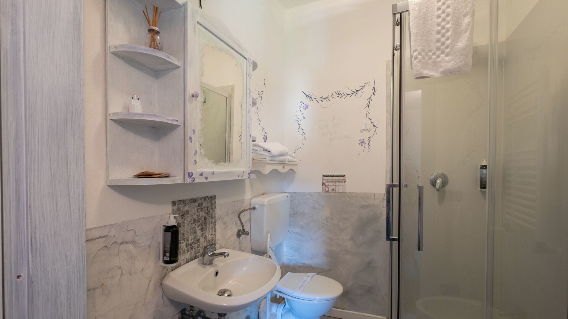 Een badkamer met een witte wastafel, een toilet, een glazen douchecabine en een spiegel met planken erboven.