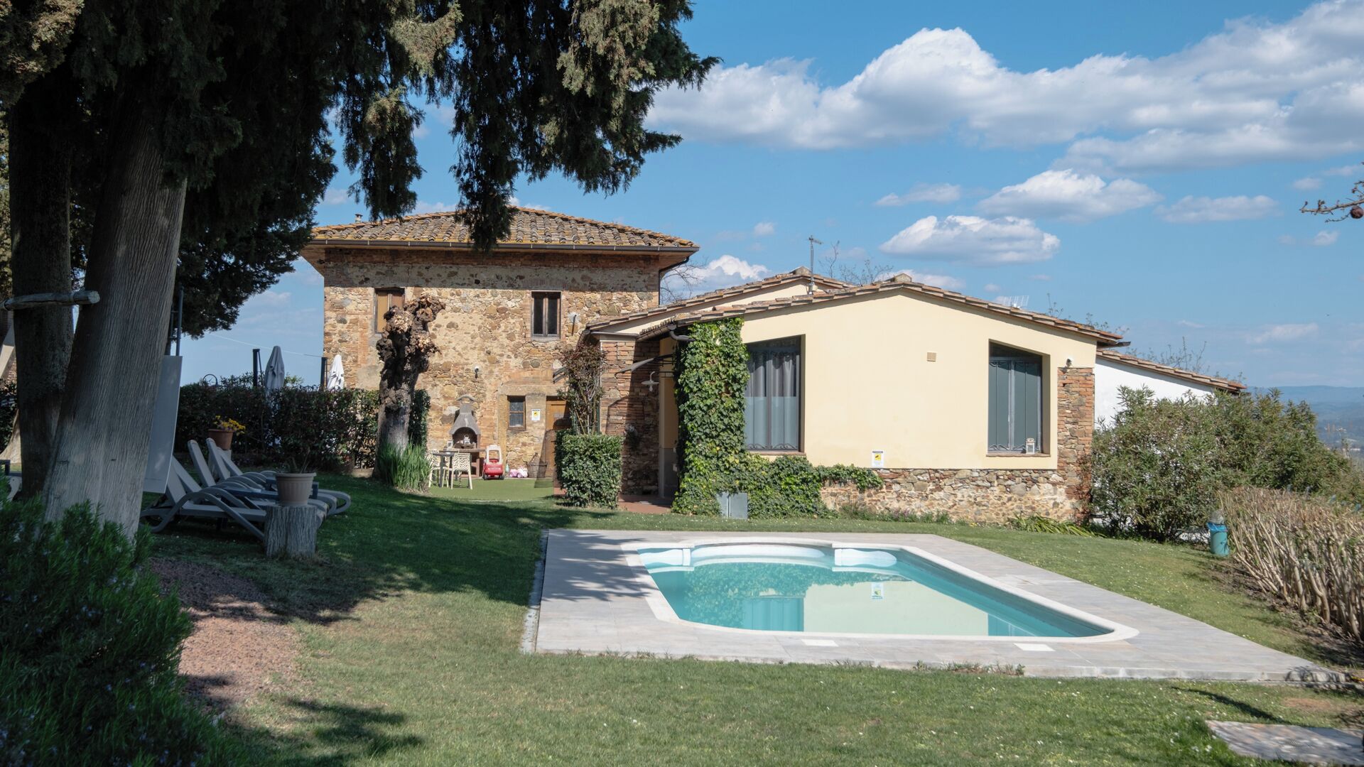A stone villa with a swimming pool, surrounded by a well-maintained garden and a large tree.