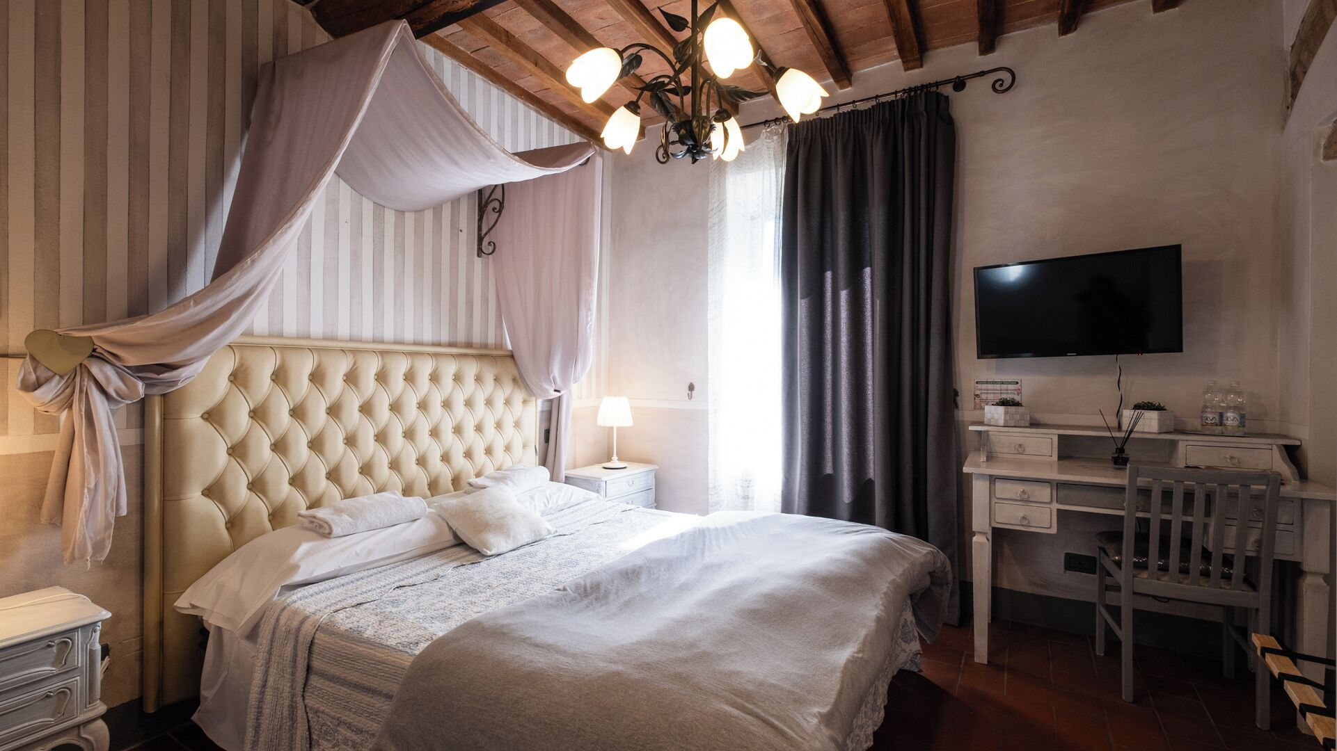 Une chambre d’hôtel avec une tête de lit capitonnée, un lit avec du linge de maison blanc, un bureau avec une chaise, un téléviseur à écran plat et une grande fenêtre avec des rideaux.