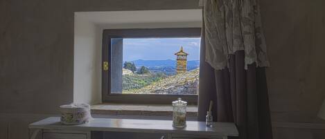 A room with a window overlooking a landscape with a tower. There is a desk with a lamp, a vase, and a container on it.